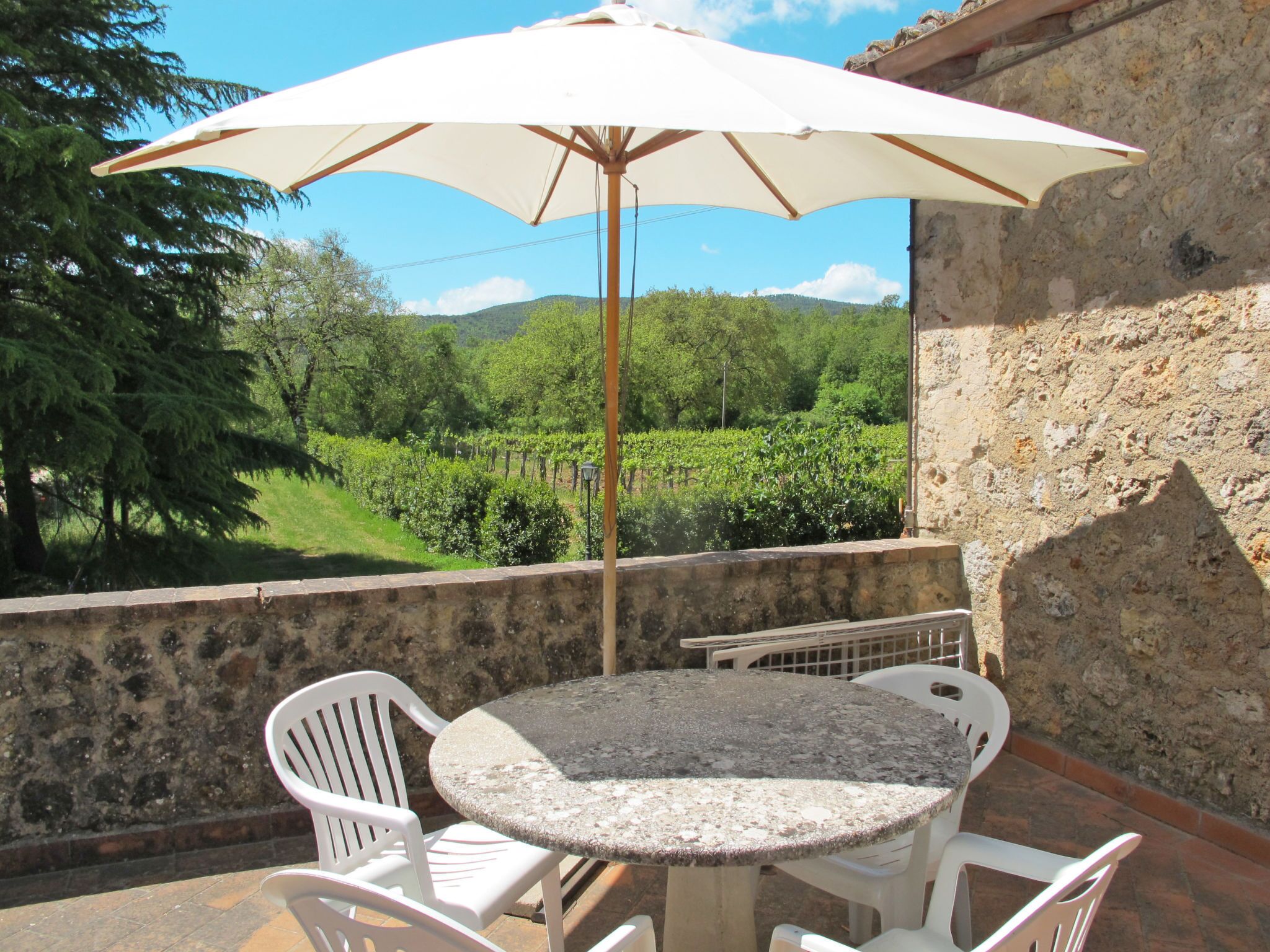 A patio with a round table and white chairs, an umbrella, and a view of a vineyard.