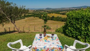 A table with a butterfly-patterned tablecloth, two white chairs, and a bottle of wine, set against a scenic backdrop of rolling hills and a clear blue sky. - Terrazza by Interhome (Istia d'Ombrone)