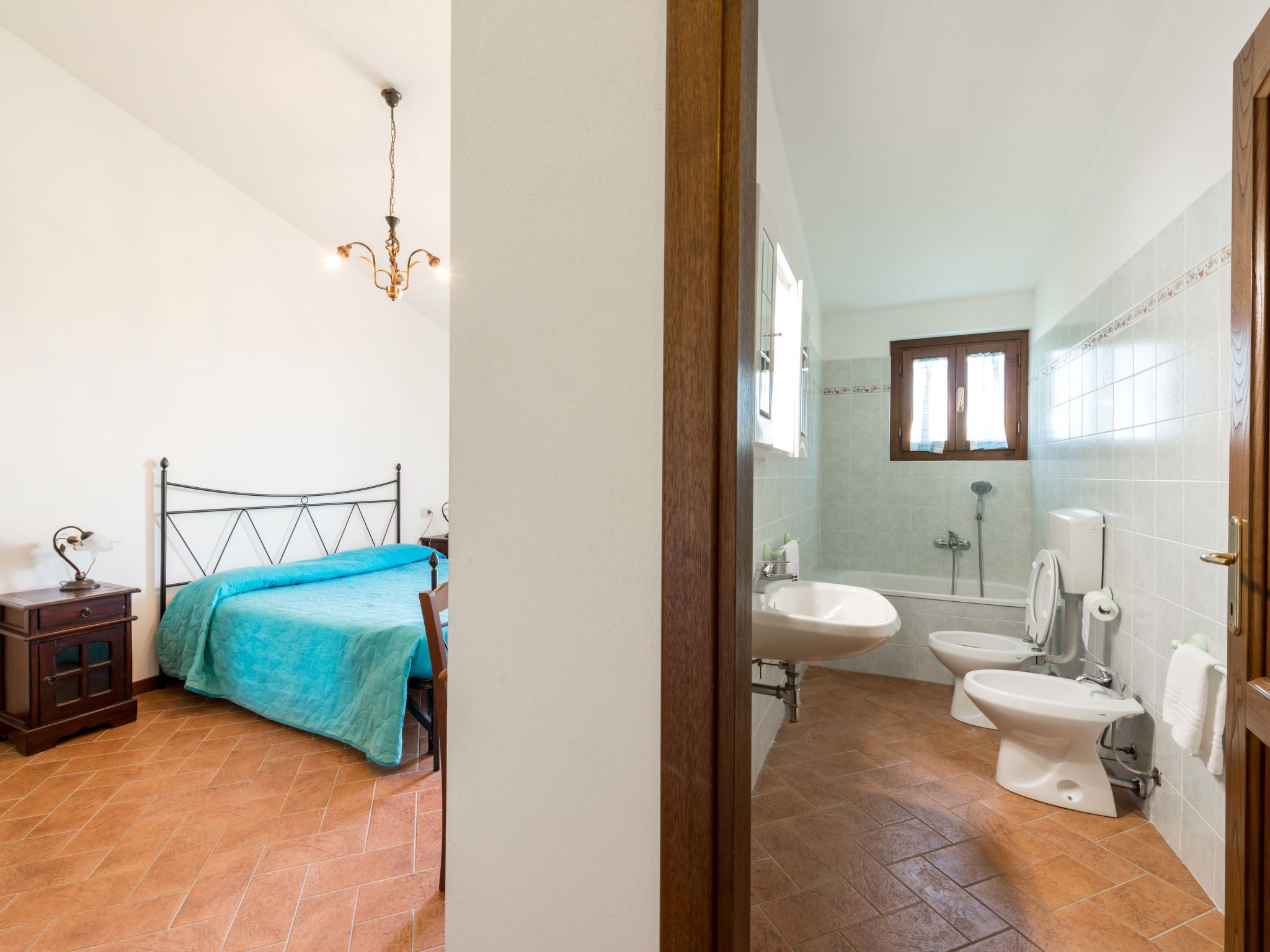 A bedroom with a bed, bedside table, and a nightstand. The room has a tiled floor and a chandelier.