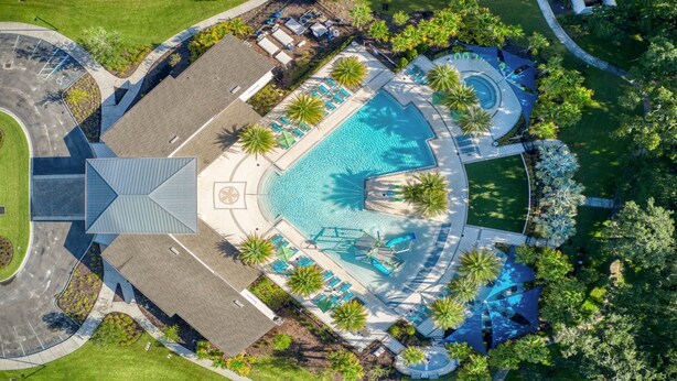 Luftaufnahme eines Resorts mit einem zentral gelegenen Swimmingpool, Liegestühlen, Palmen und einem Parkplatz.