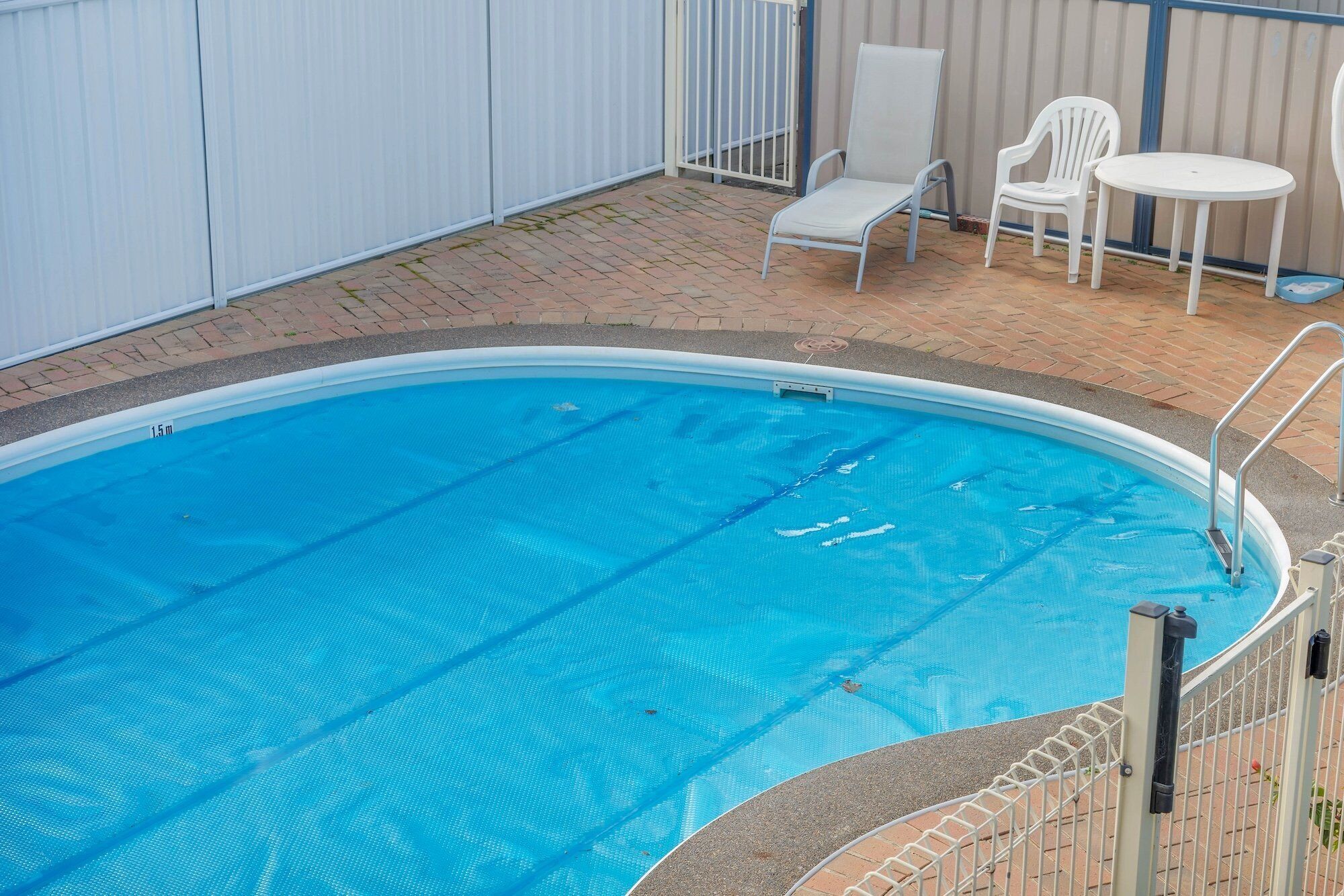 A swimming pool with a 1.5m depth marker, surrounded by a brick patio and white chairs.