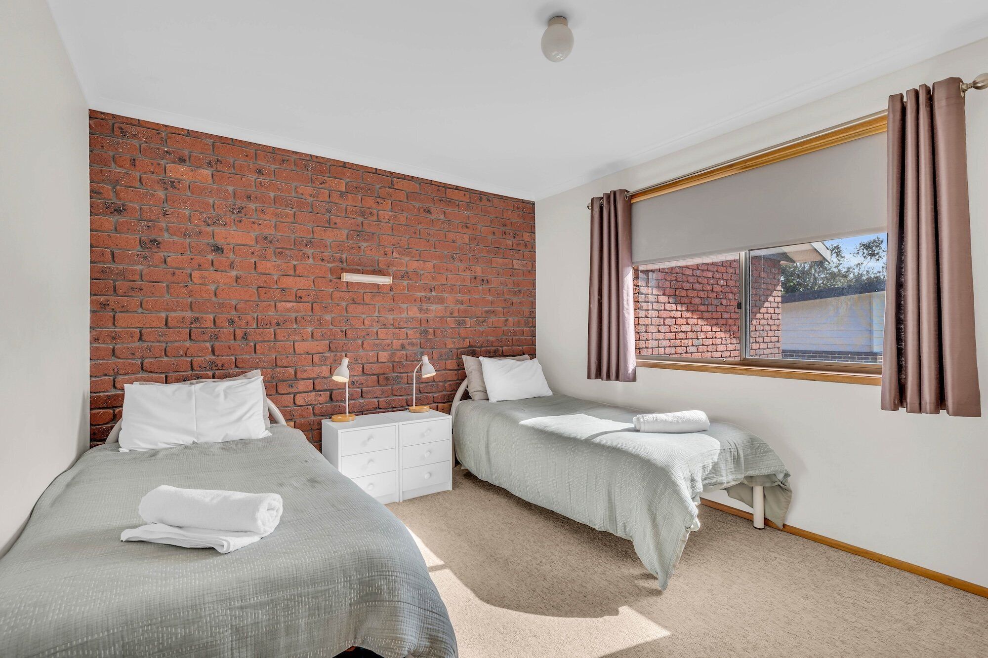 A bedroom with two beds, a brick wall, a window with curtains, and a white nightstand.