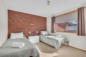 A bedroom with two beds, a brick wall, a window with curtains, and a white nightstand. - Harbour Lights - 2 Bedroom with pool (Merimbula)
