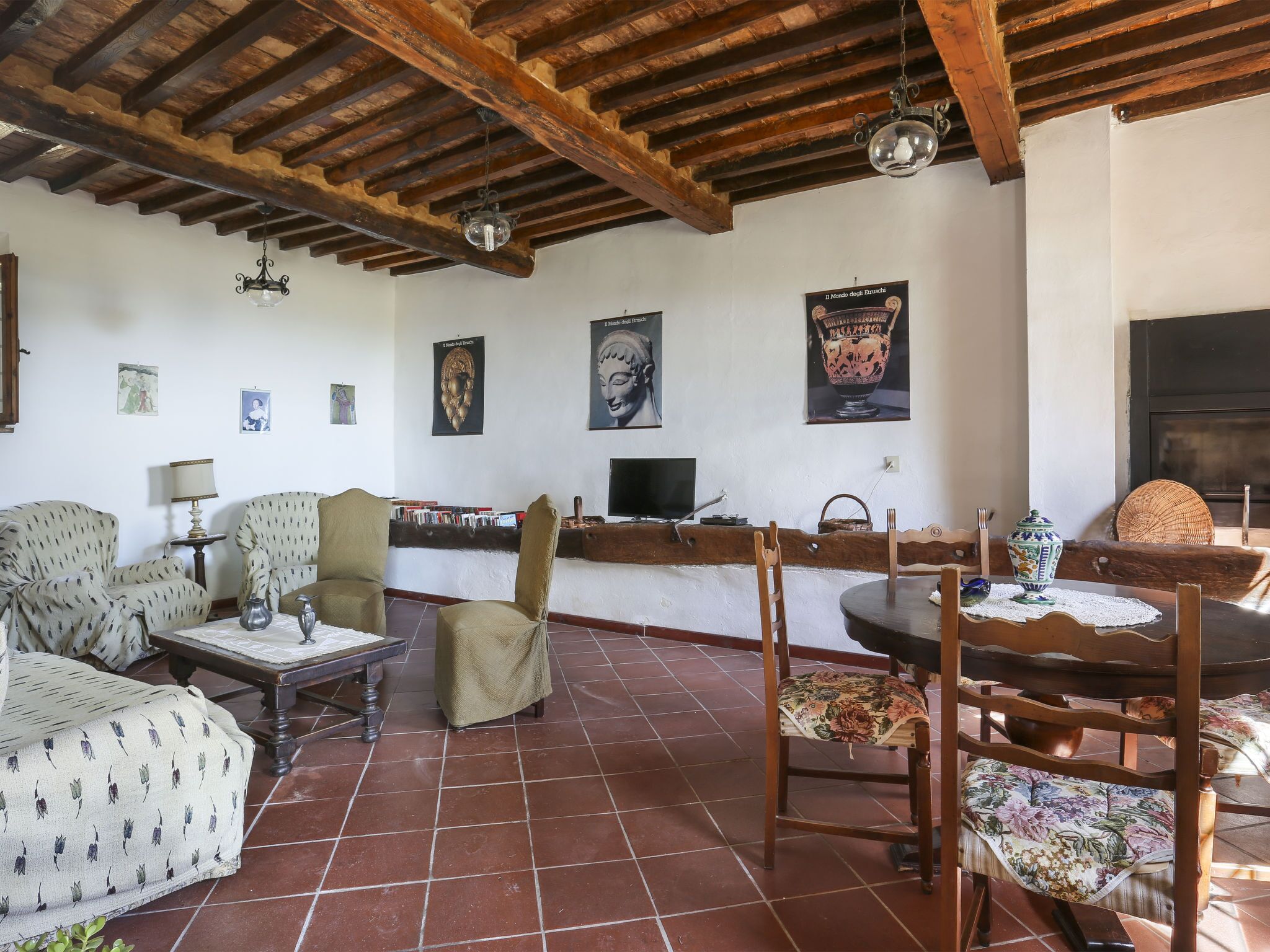 A cozy living room with wooden beams, a tiled floor, and a fireplace. There are two armchairs, a small table, and a TV on a stand.