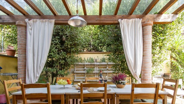 A covered patio with a wooden table and chairs, surrounded by greenery and a brick wall. - La Rondine by Interhome (San Giuliano Terme)