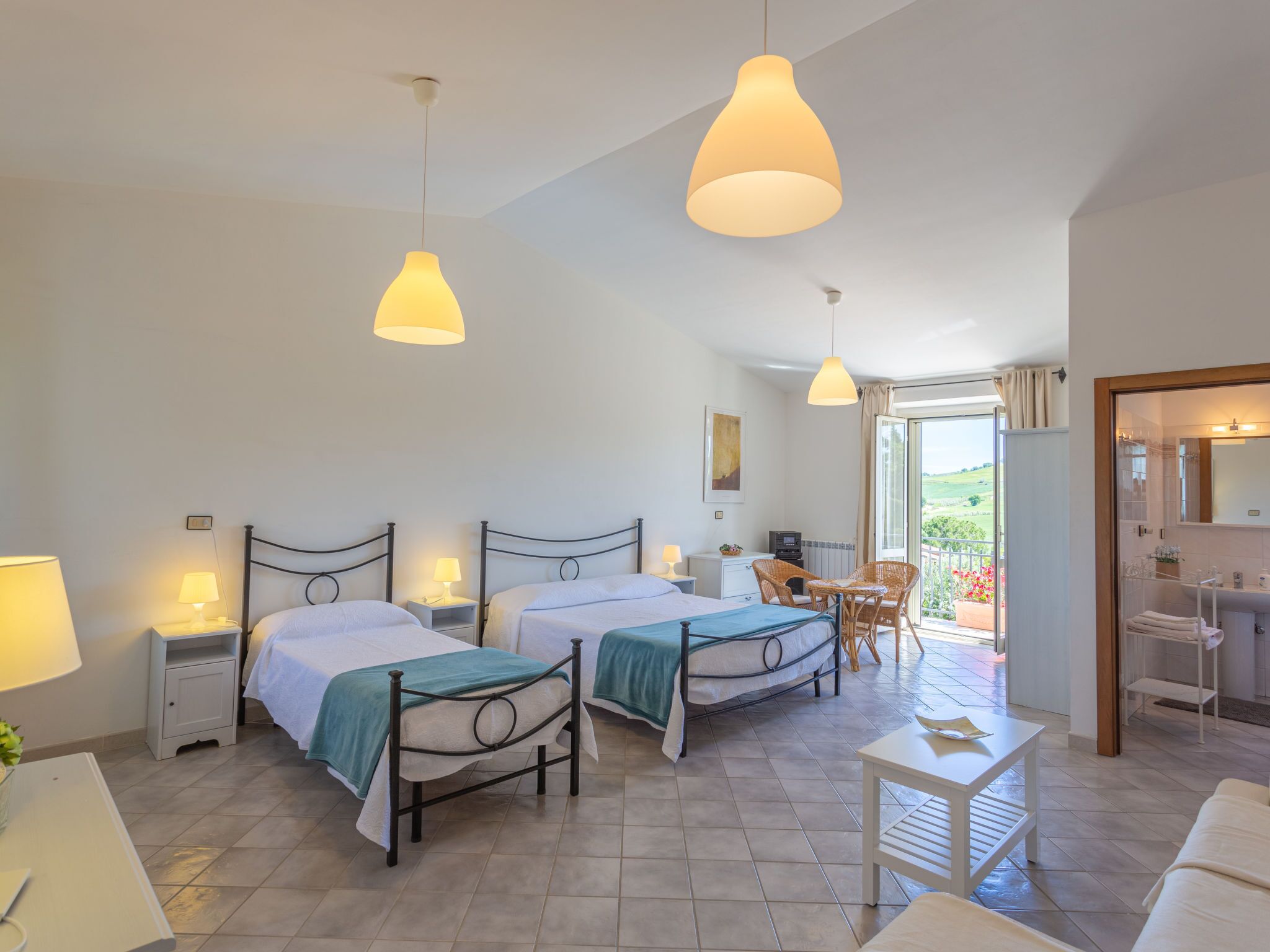 A hotel room with two beds, a small table, a chair, a balcony with a view, and a bathroom with a shower and sink.