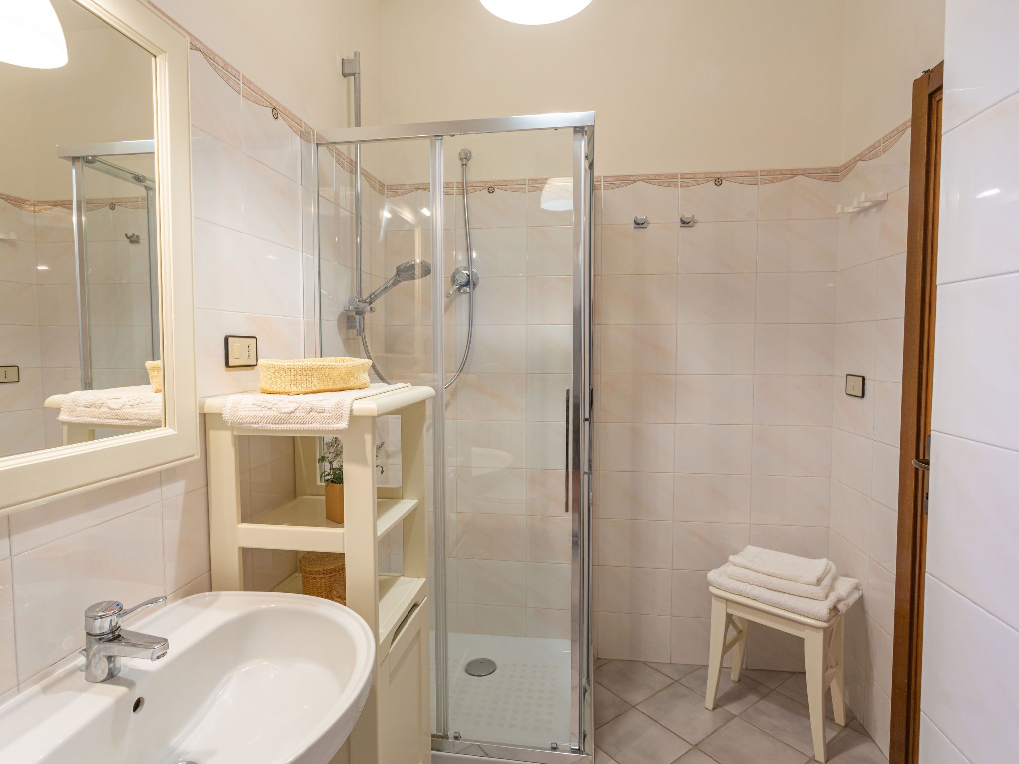 A bathroom with a glass-enclosed shower, a white sink, and a small stool with towels.