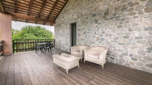 Espaço coberto ao ar livre com paredes de pedra, piso de madeira, móveis de vime e vista para o verde.