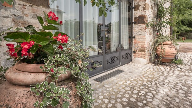 A rustic stone building with a large window, a potted plant with red flowers, and a cobblestone pathway. - Domus Magna by Interhome (Povoletto)