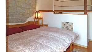 A bedroom with a stone wall, a wooden headboard, a bed with floral bedding, a nightstand with a lamp, and a wooden shelf. - Casa Polava by Interhome (Valli del Natisone)