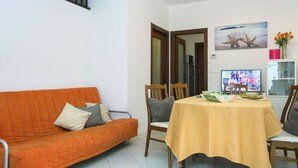Una sala de estar con un sofá naranja, una mesa de comedor con mantel amarillo y una cocina visible a través de una puerta abierta.