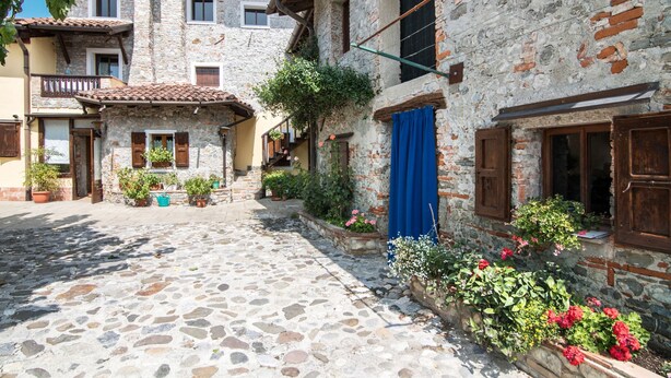 A cobblestone courtyard with stone buildings, potted plants, and a blue curtain. - Al Rustico by Interhome (San Giovanni al Natisone)