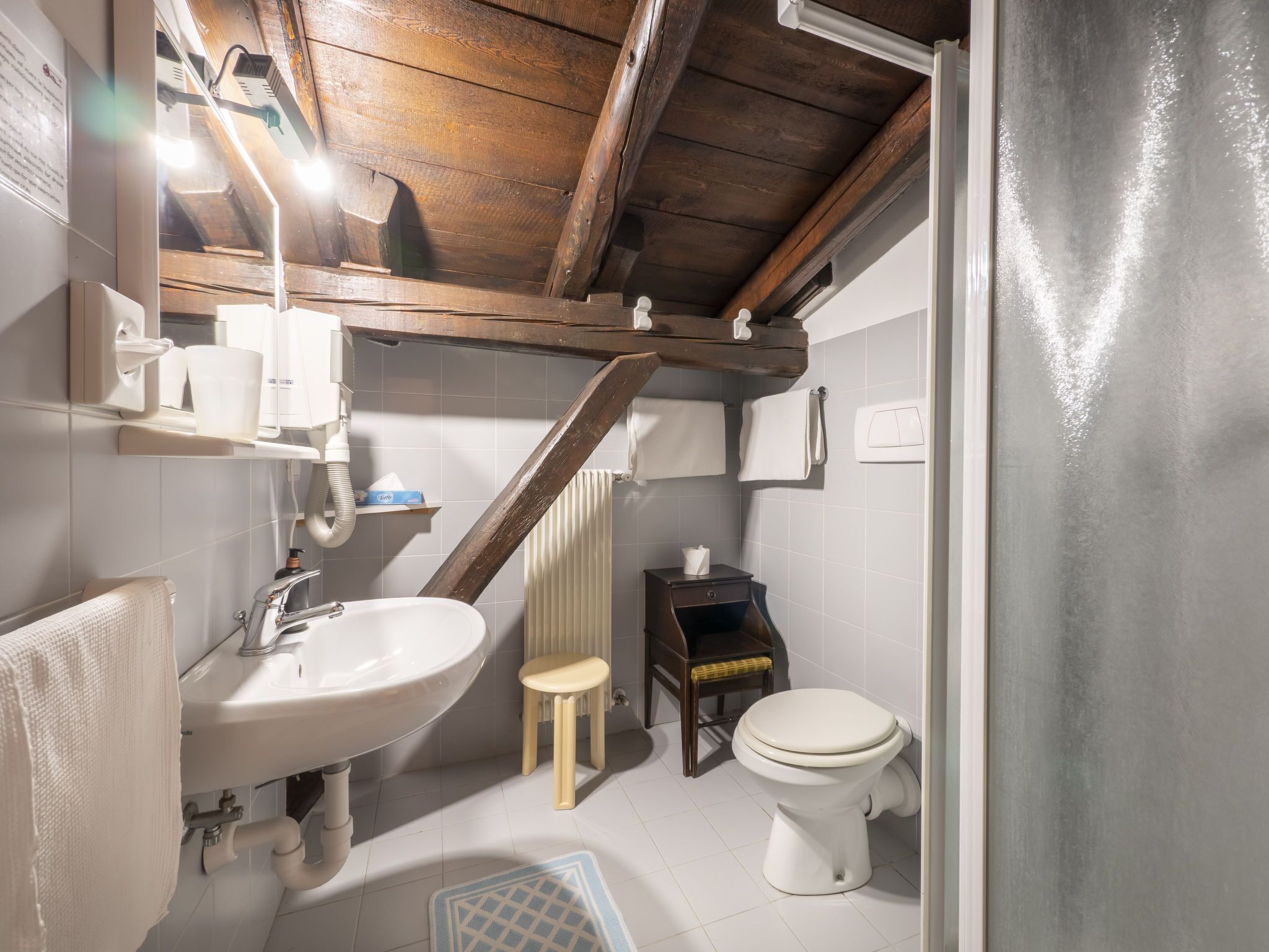 A compact bathroom with a wooden ceiling, a white sink, a toilet, and a small stool.