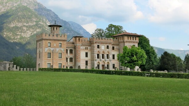 A large, historic building with multiple towers and arched windows, surrounded by a well-maintained lawn and mountains in the background.
