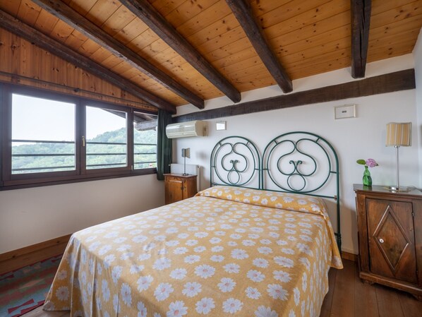 A bedroom with a bed, wooden ceiling beams, a window with a view, a radiator, a bedside table, and a cabinet.