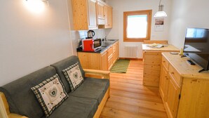 Een kleine woonruimte met een kitchenette, houten kastjes, een bank met decoratieve kussens, een televisie en een raam dat voor natuurlijk licht zorgt.