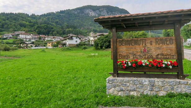 Un panneau en bois, posé sur un socle en pierre et agrémenté de jardinières, signale un espace accueillant dans un cadre rural, avec des maisons et une montagne en arrière-plan.