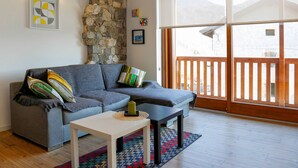 A cozy living room with a stone wall, a gray sofa, a small white table, and a view of mountains through large windows.