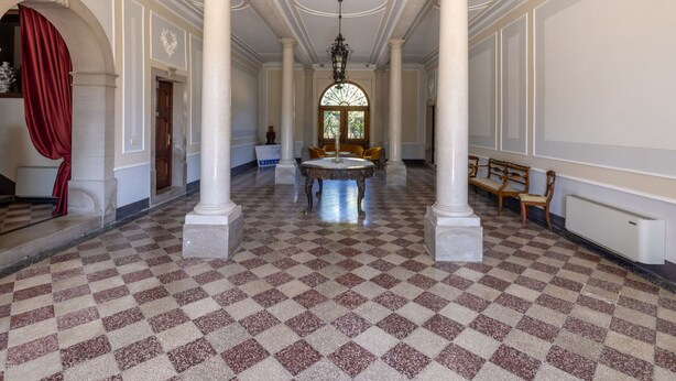 Un salón amplio con suelo a cuadros, grandes columnas y una lámpara central.