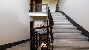 Una escalera con barandilla de madera y balaustradas de metal negro, que conduce a un rellano con una puerta y una pequeña mesa con una lámpara.