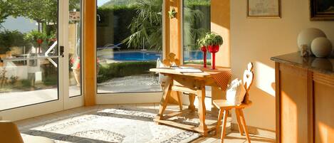 A sunlit room with a dining table, chairs, a rug, and a view of a pool and greenery outside.