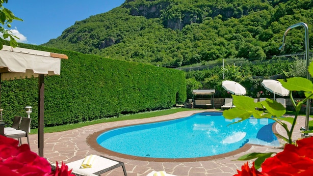 A swimming pool with lounge chairs and umbrellas, surrounded by a hedge and a mountain backdrop. - Schöneck by Interhome (Postal)