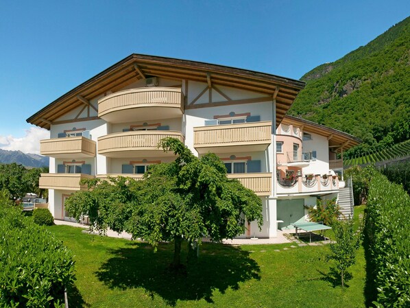 A multi-story building with balconies, surrounded by lush greenery and mountains in the background.
