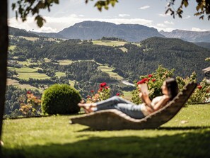 Eine Person entspannt sich in einer Hängematte mit Blick auf eine gebirgige Landschaft, einen Fluss und viel Grün.