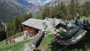Un chalet de montagne rustique, avec un toit en pierre, entouré d’une végétation luxuriante et de montagnes.