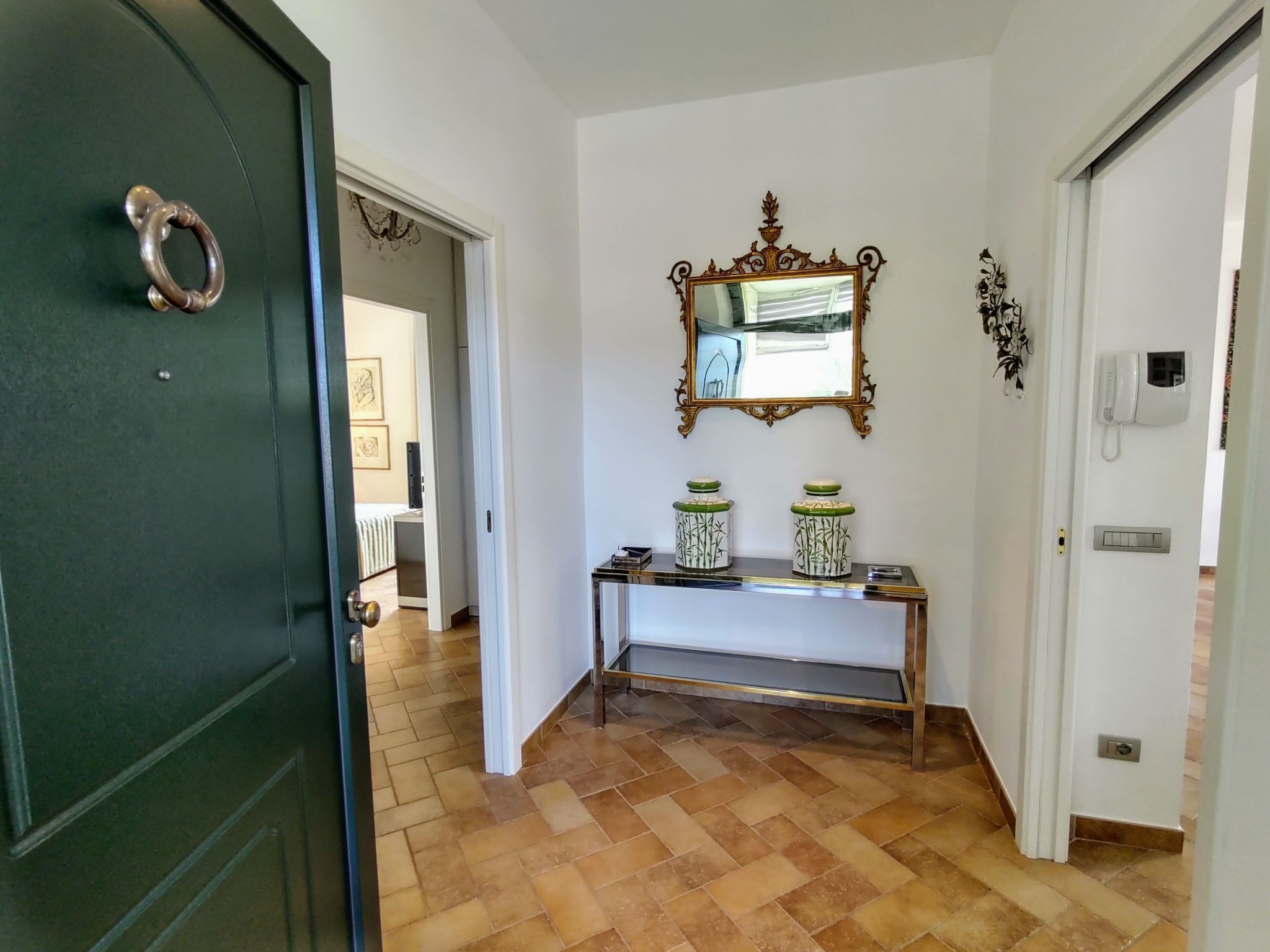 A hallway with a dark green door, a white console table with decorative jars, a mirror with an ornate frame, and a white wall with a phone and a light switch.
