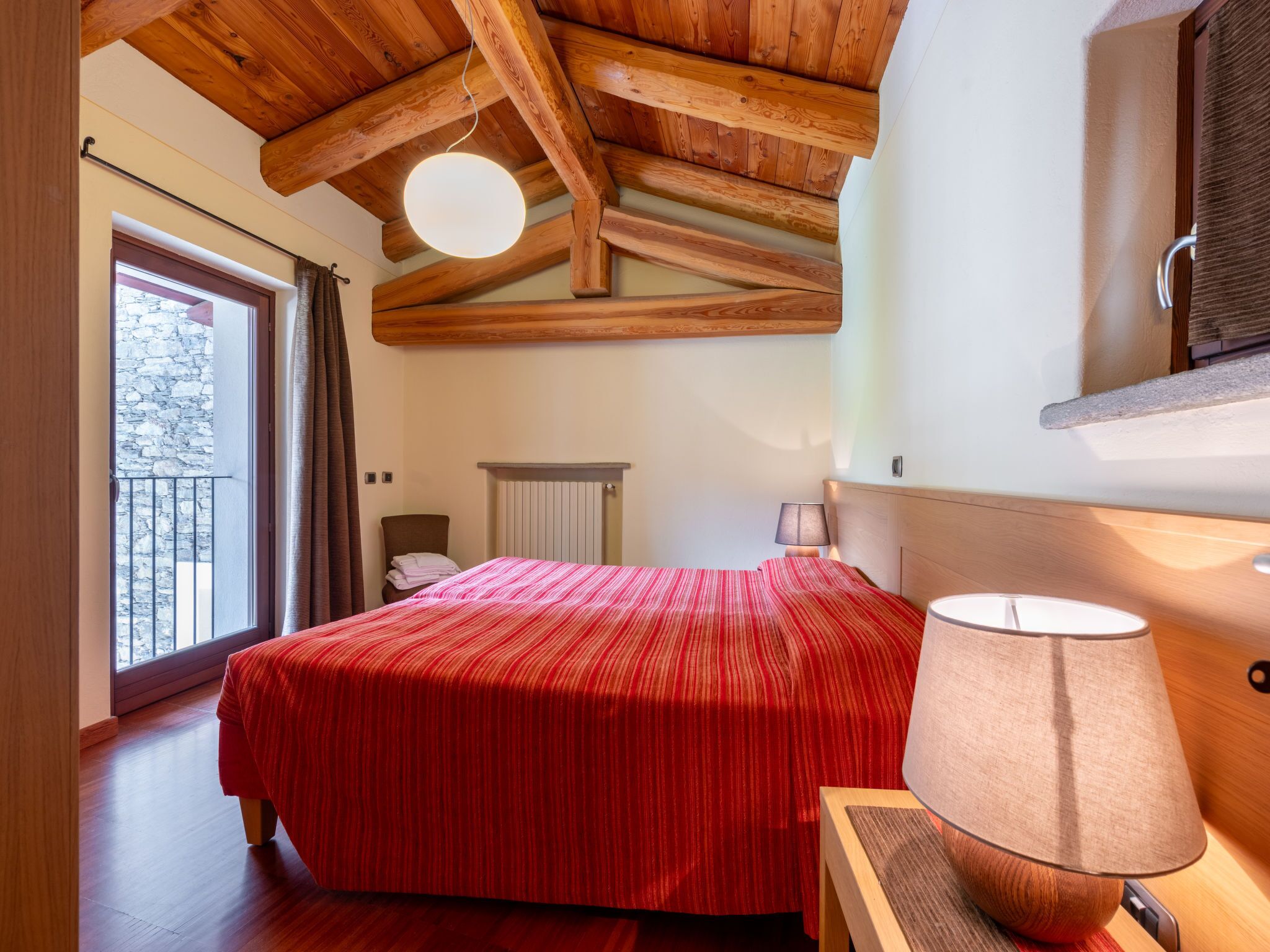A bedroom with a wooden ceiling, a bed with a red striped bedspread, a bedside lamp, and a view of a stone wall outside.