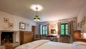 A bedroom with a large bed, a dresser, a desk, a chair, a fireplace, and a window with curtains.