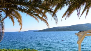 Vista sulla costa con un muro di pietra e vegetazione in primo piano, che si apre su un mare blu limpido e colline lontane.