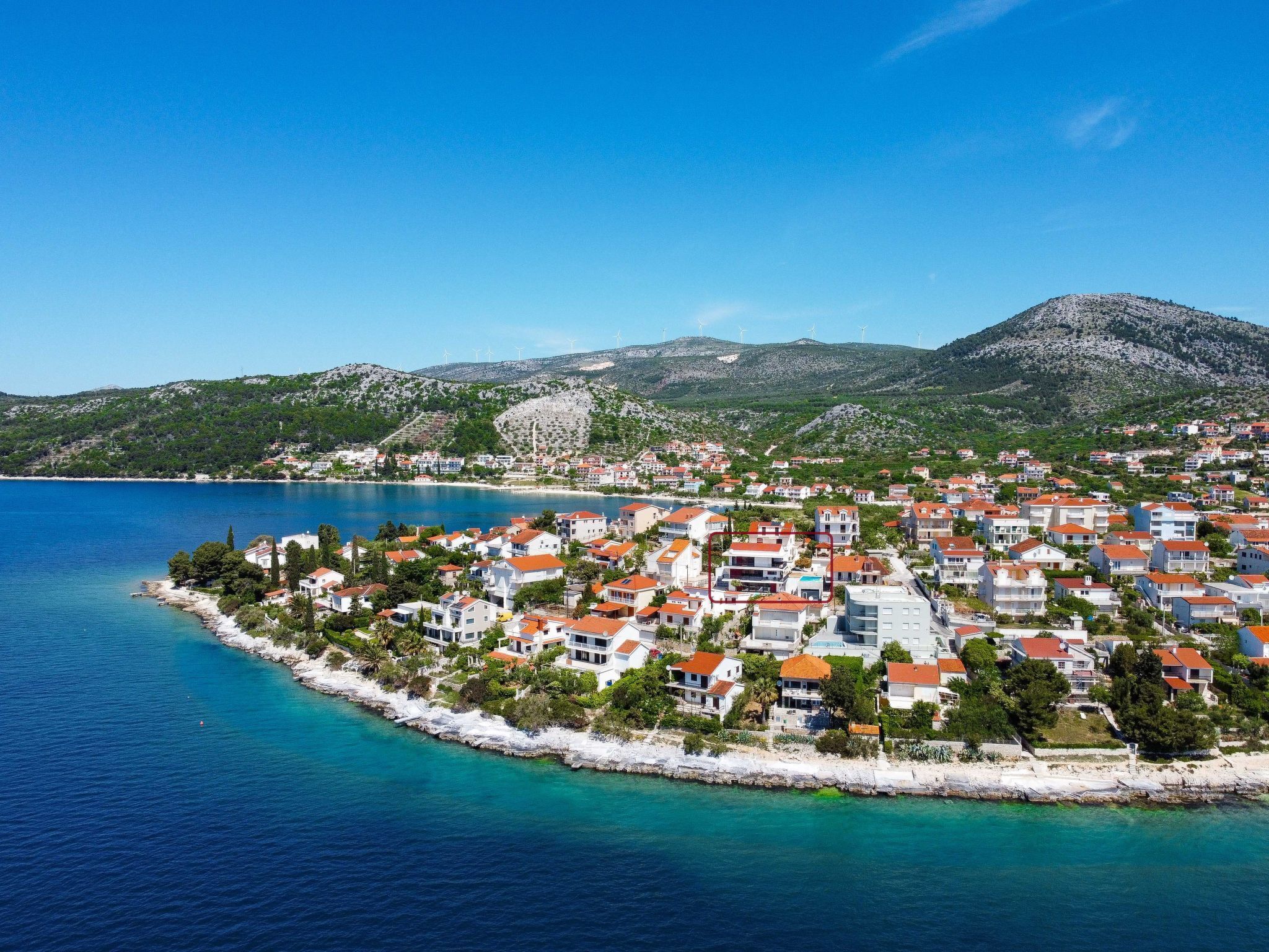 Une ville côtière avec des bâtiments qui bordent le littoral, une mer d’un bleu limpide et une chaîne de montagnes en arrière-plan.
