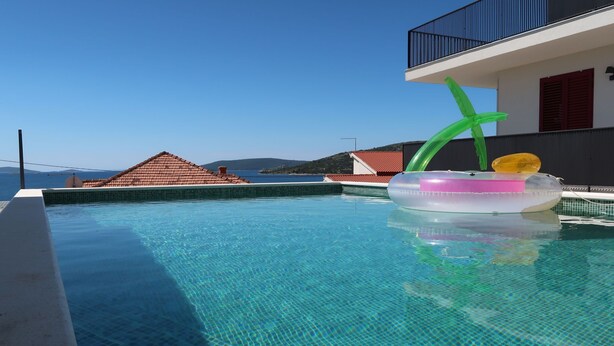 Una piscina blu cristallina con un galleggiante gonfiabile verde, un solarium piastrellato e vista su edifici con tetti rossi.
