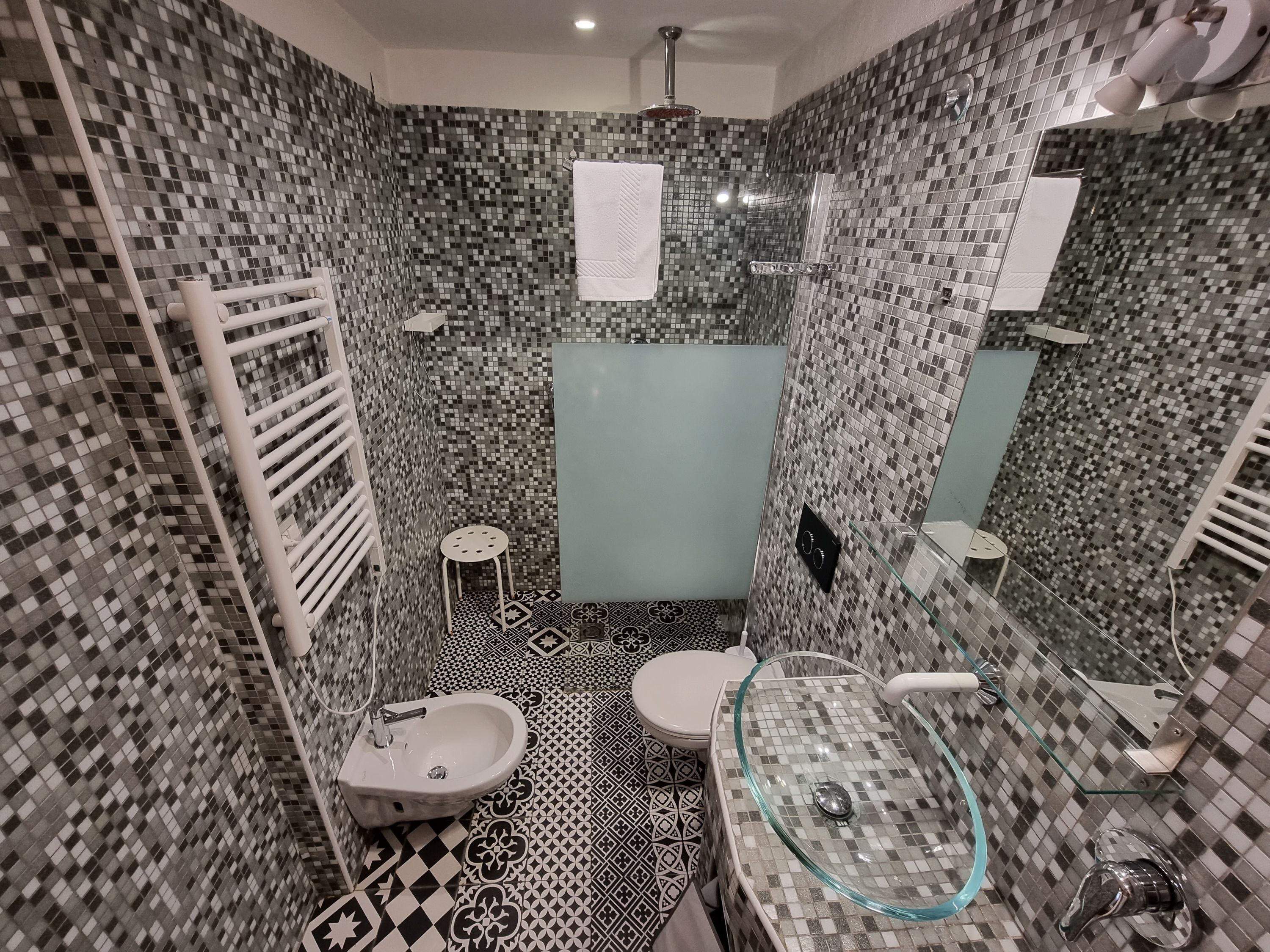 A bathroom with mosaic tile walls, a glass-enclosed shower, a pedestal sink, and a toilet.