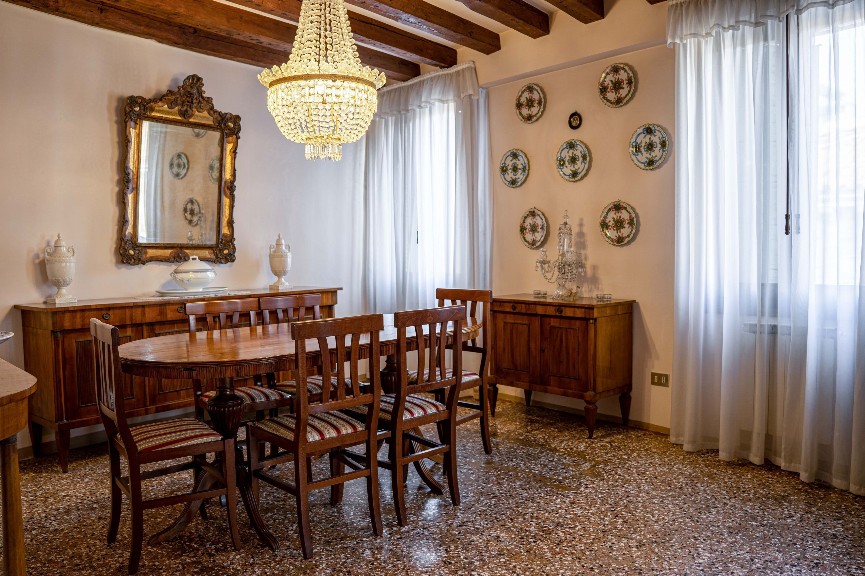 Een eetkamer met een houten tafel en stoelen, een kroonluchter, een spiegel en decoratieve borden aan de muur.