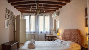 A hotel room with a large bed, a wooden headboard, a chandelier, a desk with a lamp, and a window with curtains. - Casa Allegra by Ca-sa Check (Venice)