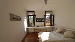 A bedroom with a bed, a desk with stools, a window with an open shutter, and a ceiling fan. - Apartment in the Heart of Venice by Ca-sa Check (Venice)