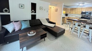 A modern living room with a dark sectional sofa, a small coffee table, and a kitchen area with wooden cabinets and a microwave. - Pleasant Mountain Escape 5BR Condo with Moose Pond Access (Bridgton)