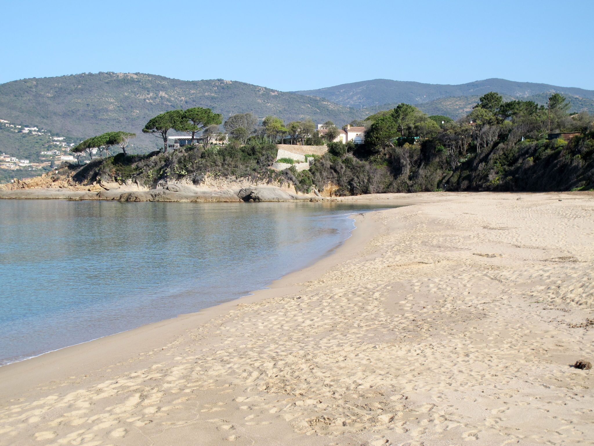 Una playa con agua cristalina, orilla de arena y casas al fondo.