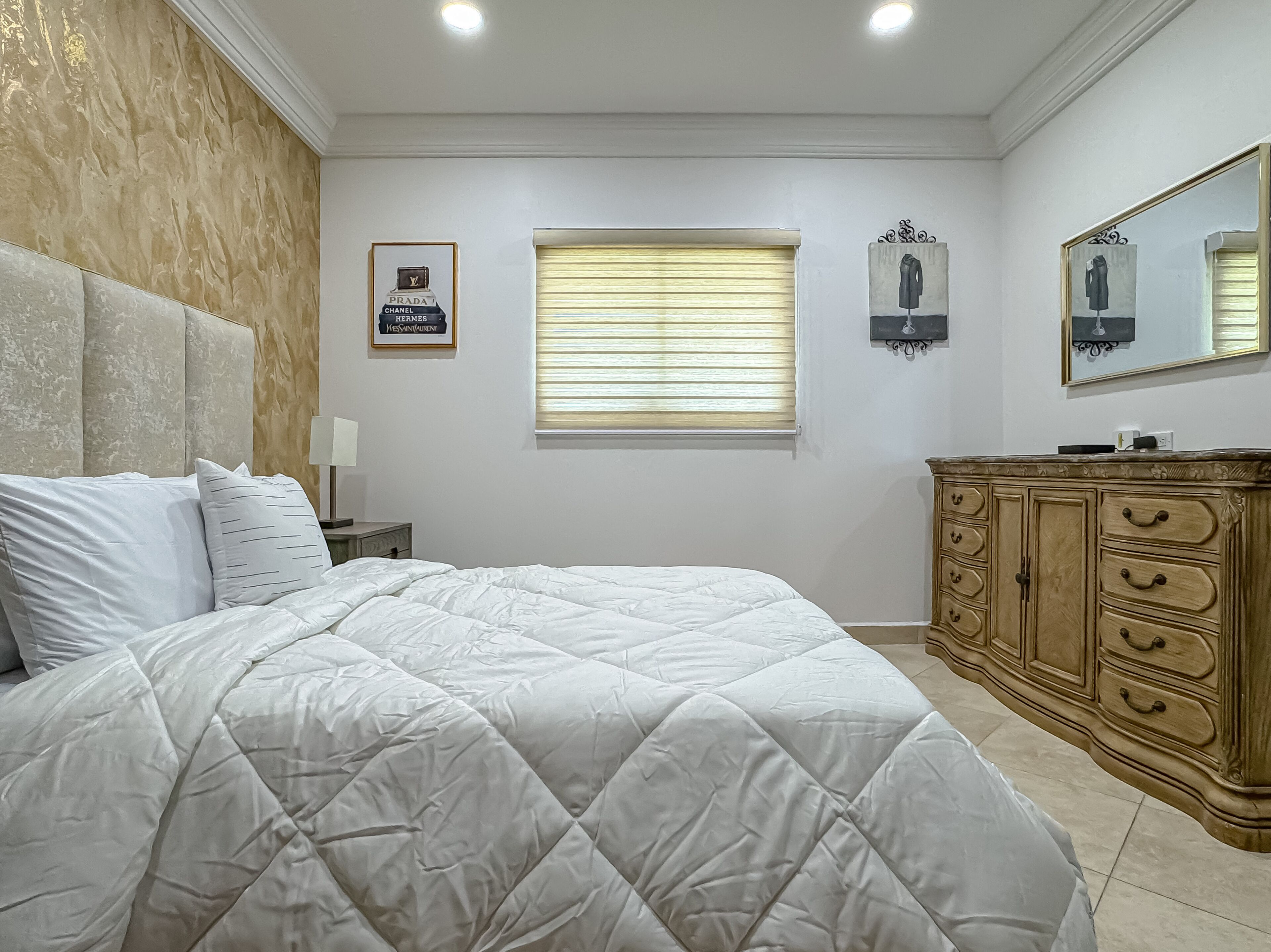 A bedroom with a large bed, a wooden dresser, a framed picture of a perfume bottle, and a window with blinds.
