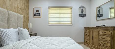 A bedroom with a large bed, a wooden dresser, a framed picture of a perfume bottle, and a window with blinds.