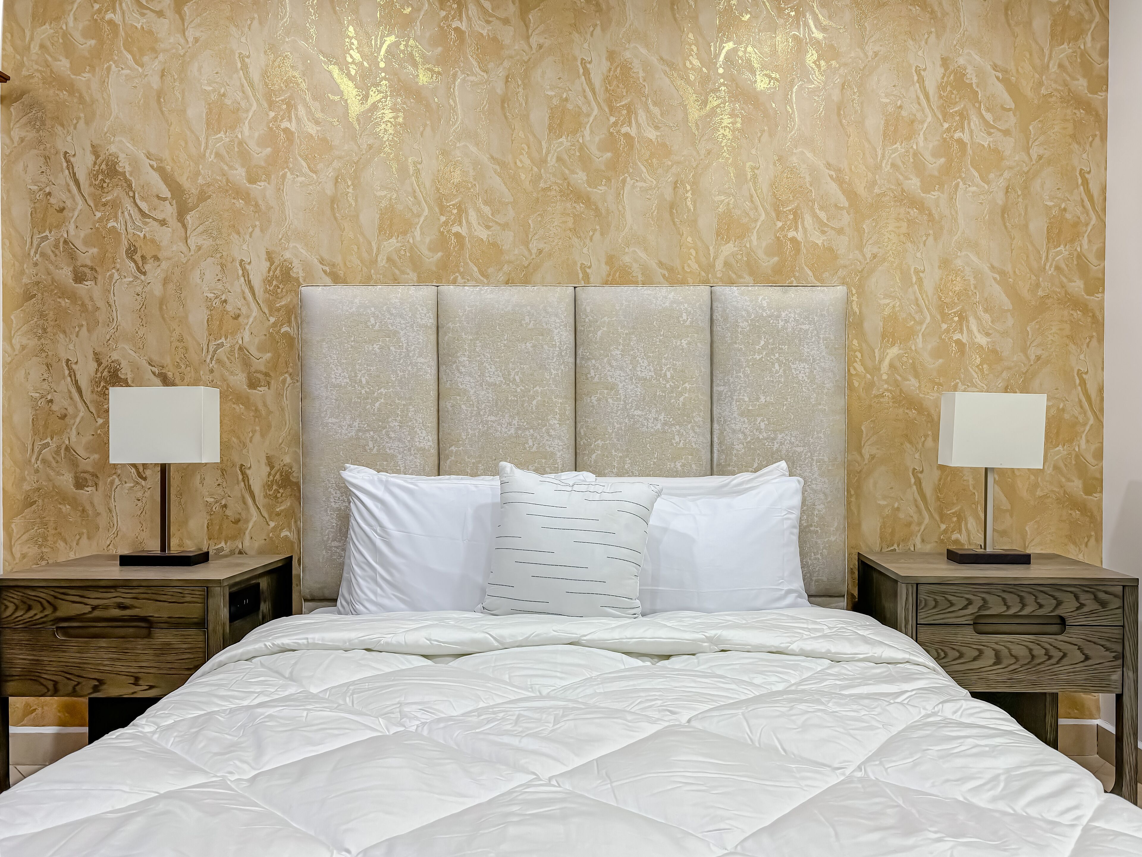 Camera da letto con testiera effetto marmo, copriletto bianco e due comodini con lampade.