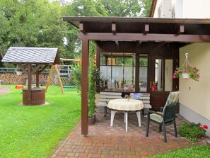 Un patio couvert avec une balançoire en bois, une petite table avec une chaise et une jardinière suspendue.