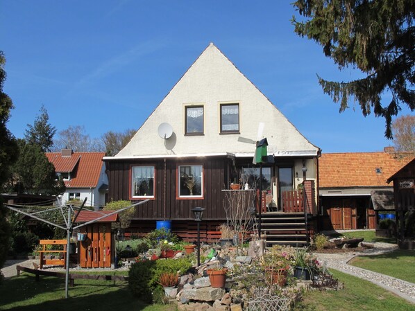 A two-story house with a sloped roof, a satellite dish, and a small garden with various plants and a wooden bench.