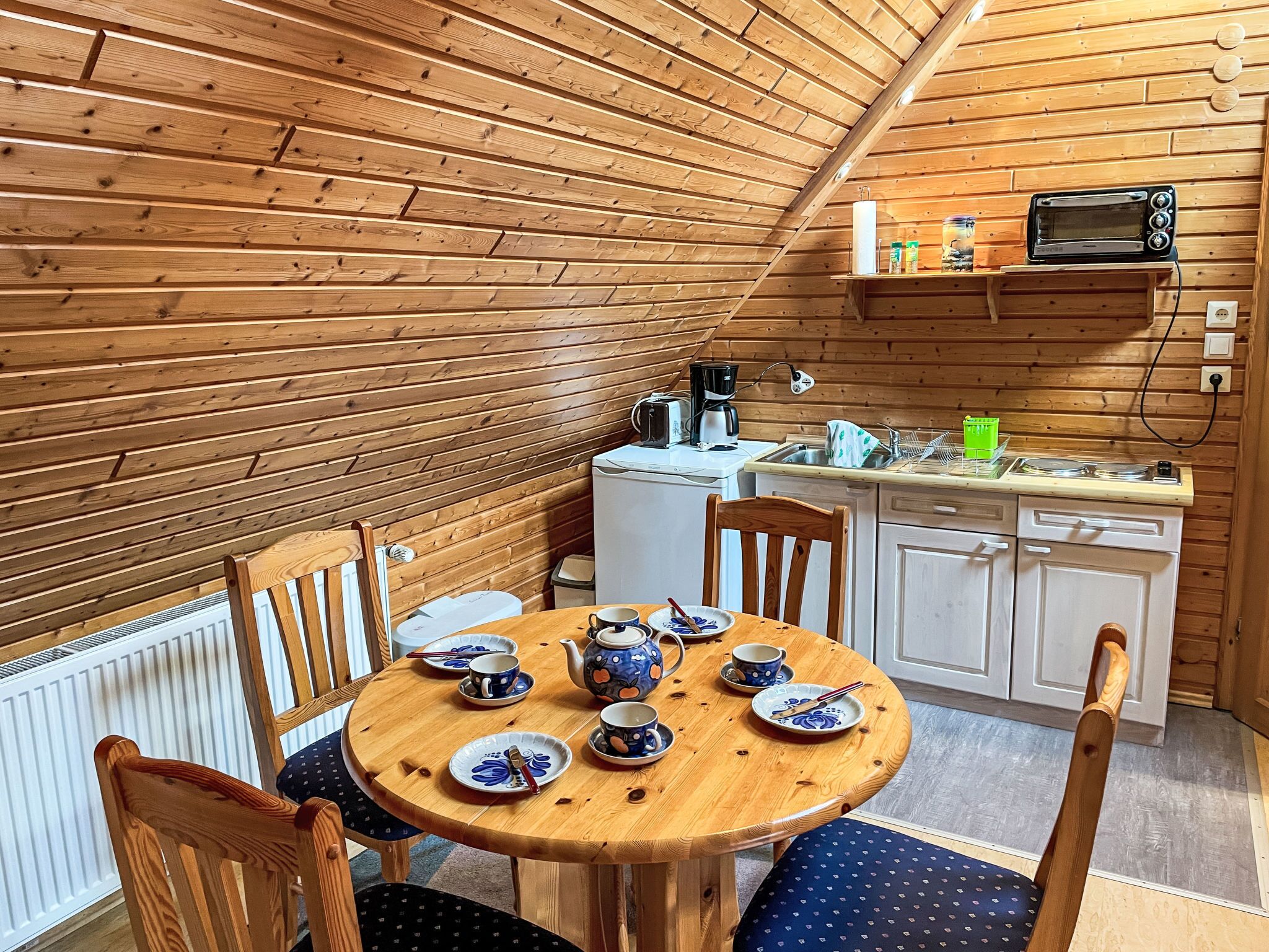 A cozy wooden cabin with a dining area featuring a round wooden table set with plates, cups, and a teapot. There is a kitchen area with a microwave, sink, and cabinets.