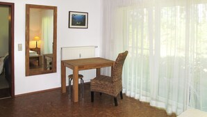 Ein Hotelzimmer mit Holztisch, Korbstuhl, großem Spiegel, gerahmtem Bild an der Wand und Fenster mit durchsichtigen Vorhängen.