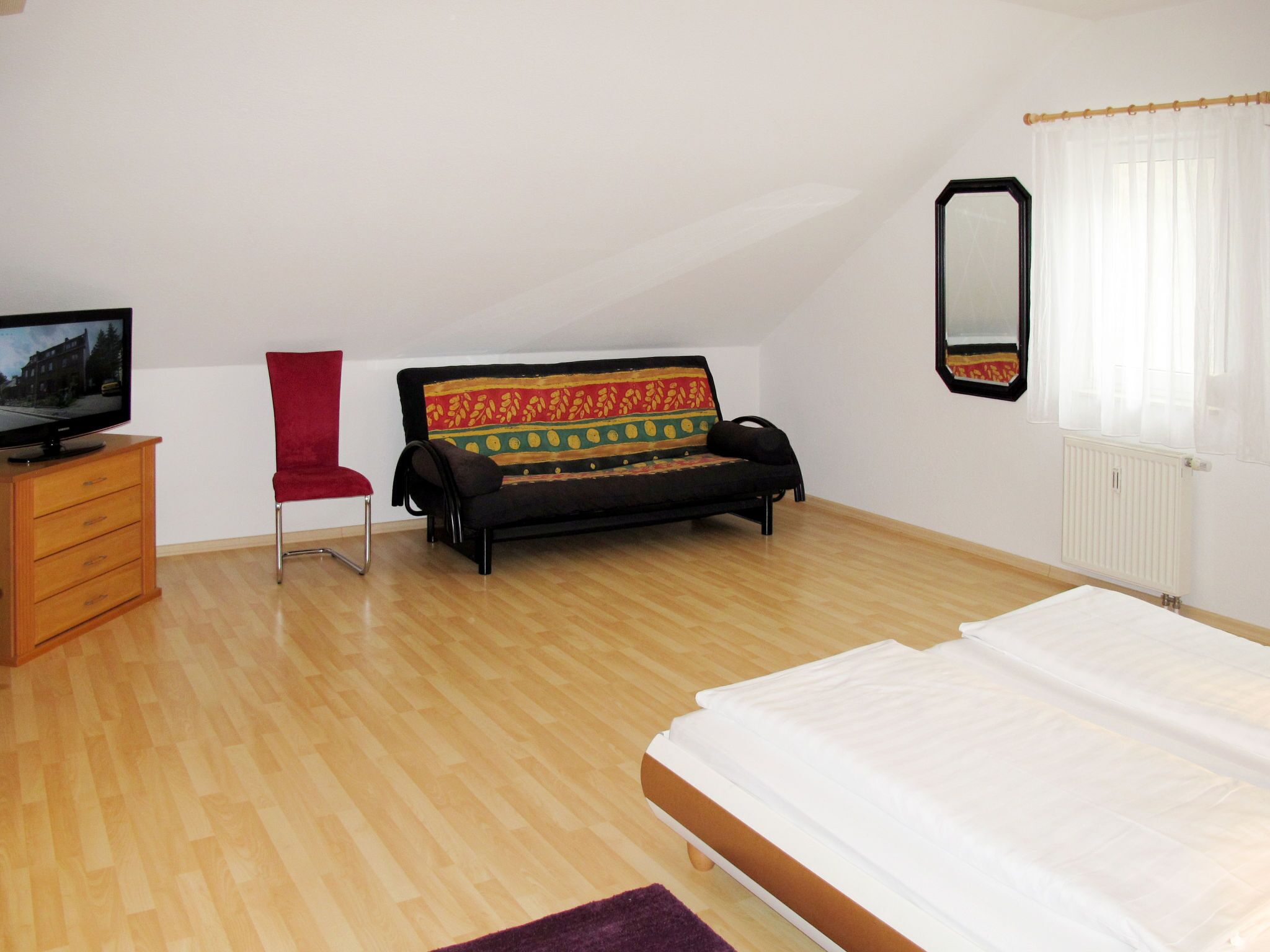 A bedroom with a bed, a sofa, a TV on a wooden cabinet, a red chair, and a window with curtains.