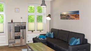 A living room with a dark grey sofa, a wooden coffee table, and a blue rug. There is a window with blinds, a radiator, and a picture on the wall. - Apartment Helene by Interhome (Ribnitz-Damgarten)